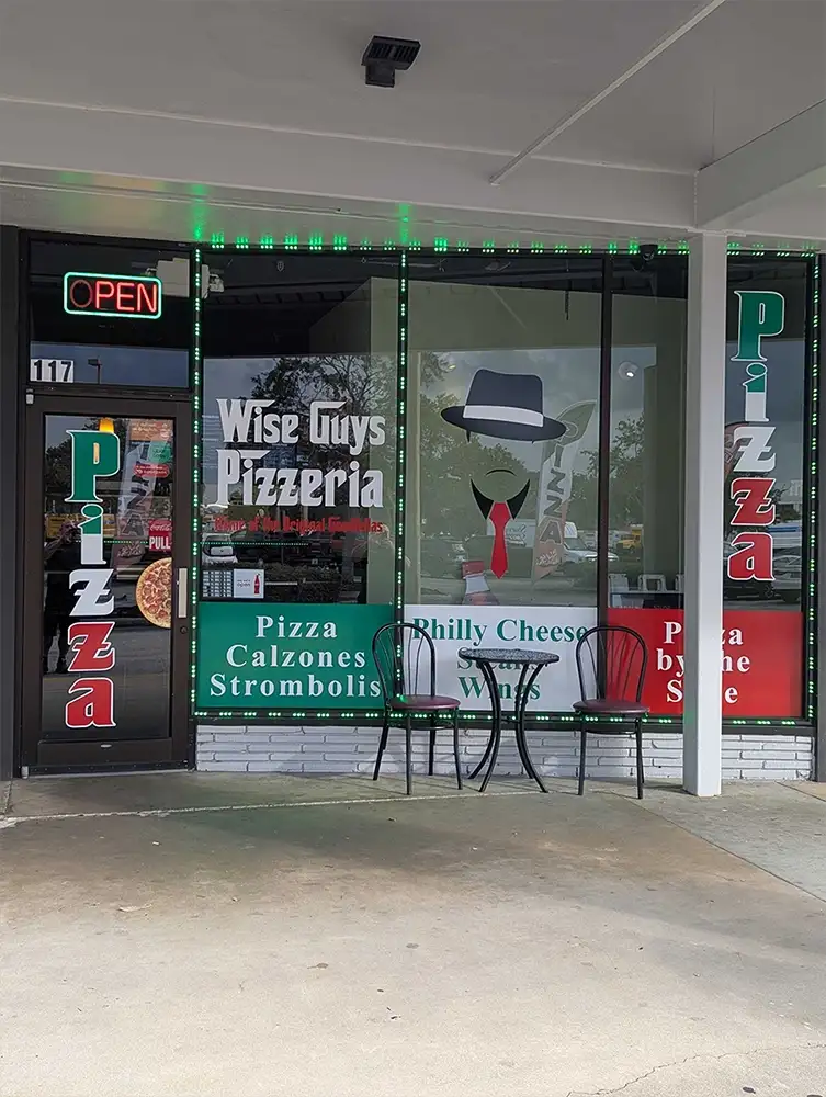 Exterior of Wise Guys Pizzeria in Florida, featuring neon open sign, pizza and calzone menu items, a stylized character